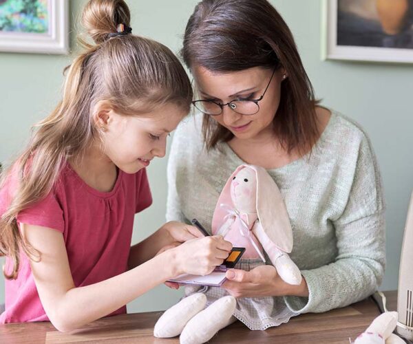 Mother and daughter child play together at home, girl and mom make make-up paints cheeks to toy handmade doll bunny sewn on sewing machine
